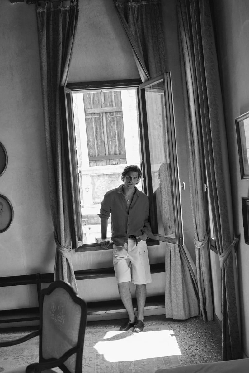 Black-and-white interior image of model Tony Özkan standing in front of an open window in a linen shirt and tailored shorts, framed by curtains and Venetian shutters