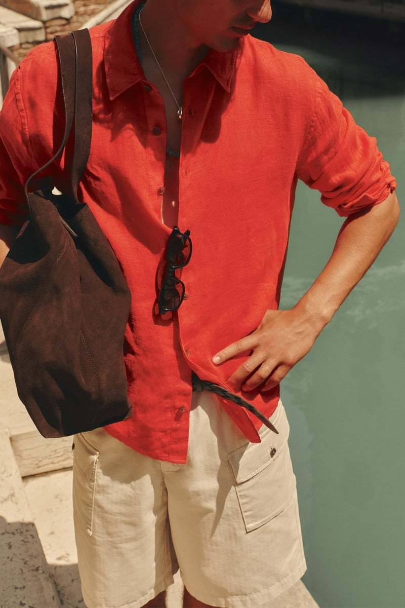 Close-up of model Tony Özkan in a bright red linen button-down shirt tucked into light cargo shorts, with sunglasses hooked at the chest and a brown suede tote slung over the shoulder