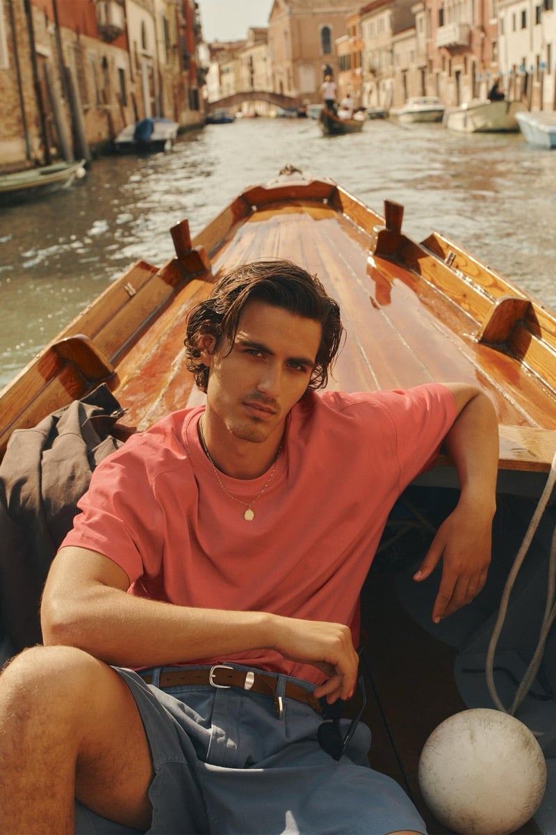 Model Tony Özkan reclining in a wooden boat drifting along a Venetian canal, wearing a coral pink crewneck tee and slate blue belted shorts