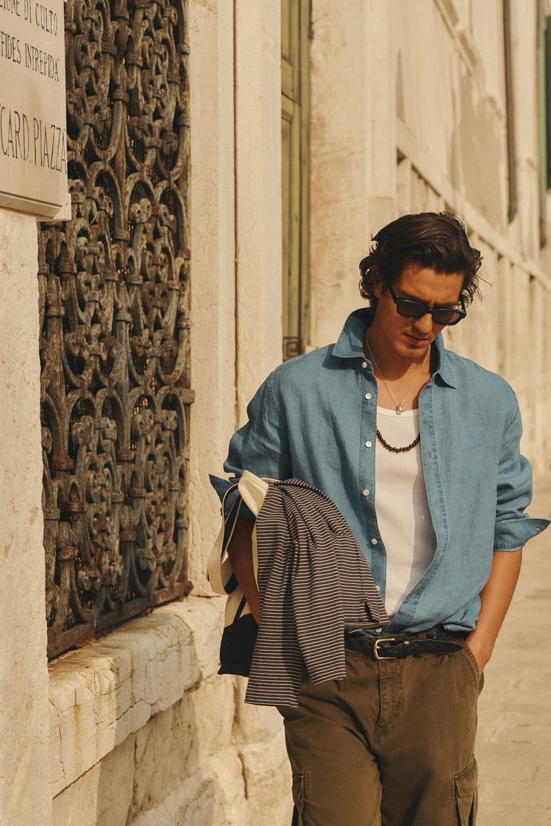 Model Tony walking a Venetian street in a blue linen shirt worn open over a ribbed tank with olive cargo trousers and sunglasses, holding a striped overshirt and a tote bag