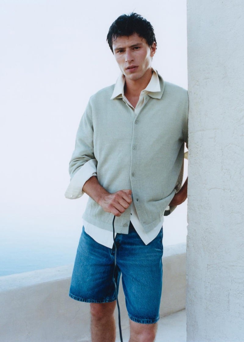 Clément Laguardia leans against a whitewashed wall in a knit cardigan layered over a linen shirt with mid-length blue denim shorts