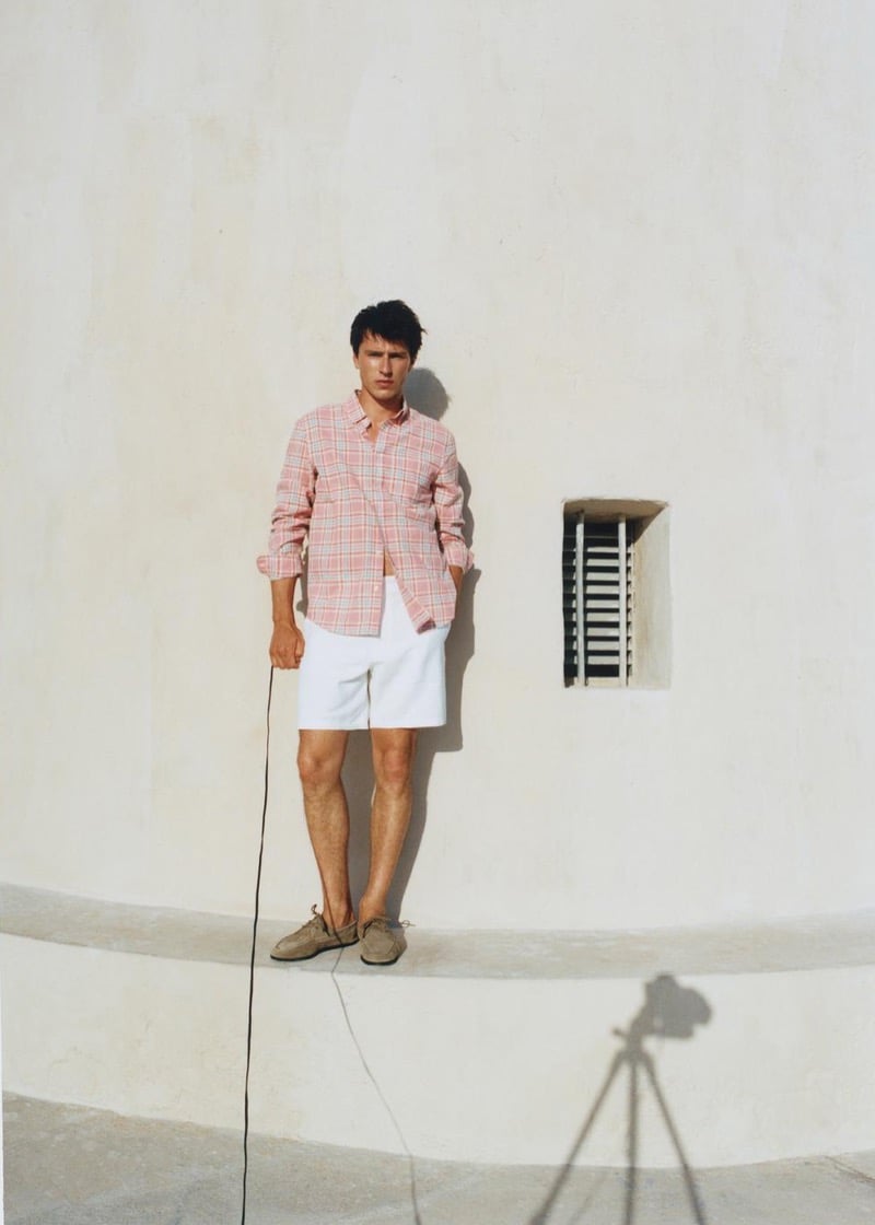 Clément Laguardia stands on a curved white stucco ledge in a pink plaid button-up shirt and white shorts with tan boat shoes