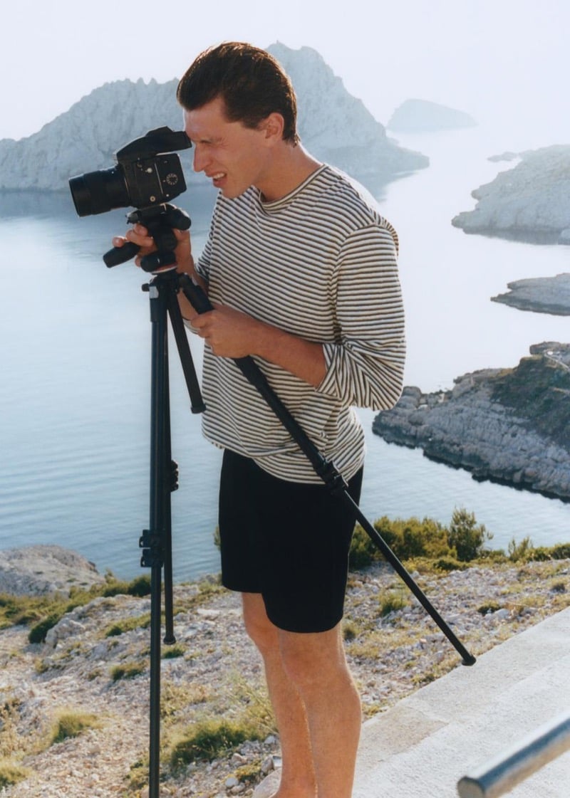 Clément Laguardia looks through a film camera mounted on a tripod above a Mediterranean cove, wearing a striped long-sleeve top with black shorts