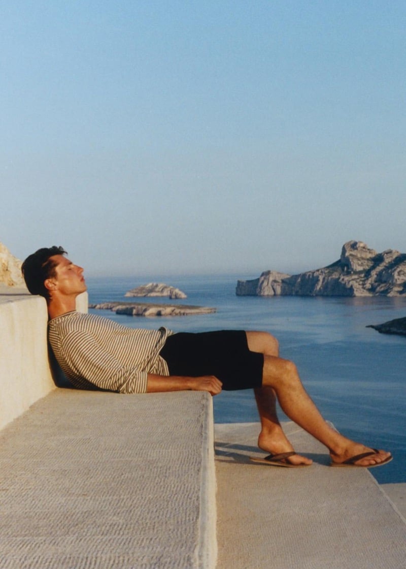 Clément Laguardia reclines on a stone ledge above the sea in a striped long-sleeve top, black shorts, and brown flip-flops, his face tilted toward the sun