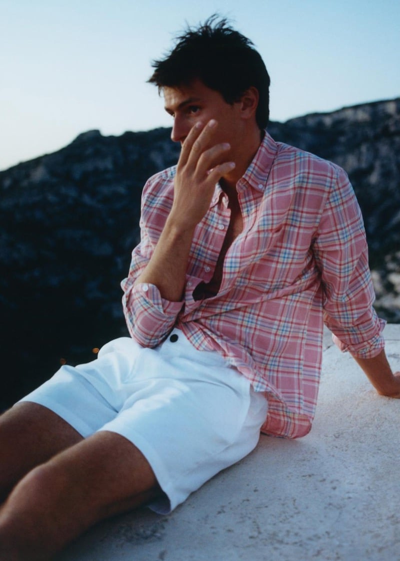 Clément Laguardia sits on a sunlit ledge in a pink plaid shirt and white shorts, his hand raised to his face with rocky cliffs behind him