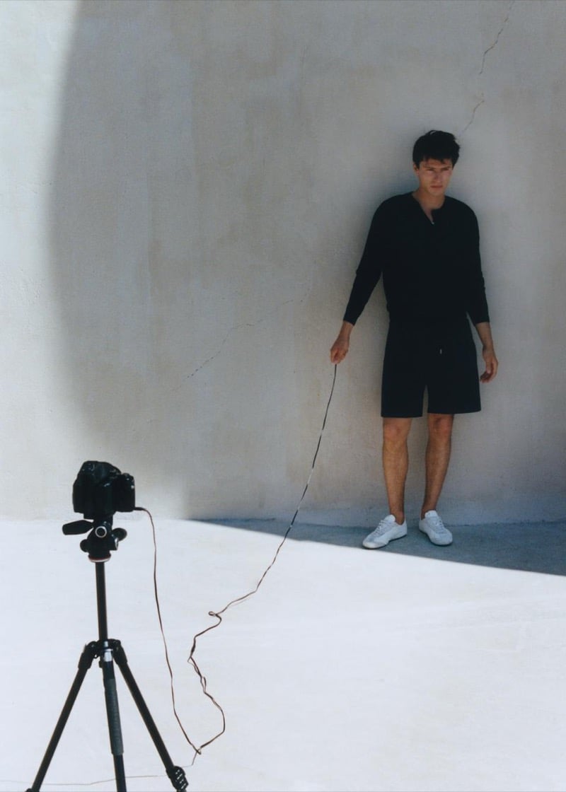 Clément Laguardia stands against a cracked stucco wall in a black henley sweatshirt and matching shorts with white sneakers, holding a remote shutter cable to the tripod-mounted camera opposite him