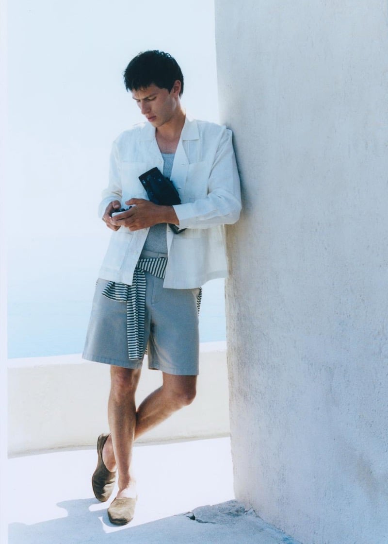 Clément Laguardia leans against a whitewashed corner in a white linen camp-collar overshirt, a gray tee and shorts, and a striped knit knotted at the waist