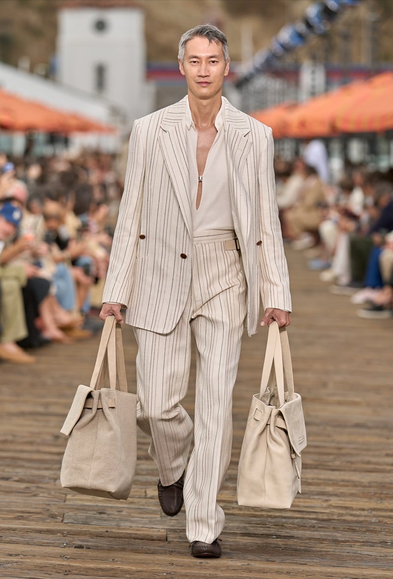 Male model walks the Malibu Pier runway in a cream pinstripe double-breasted suit worn over a matching pale knit polo, carrying two oversized canvas totes