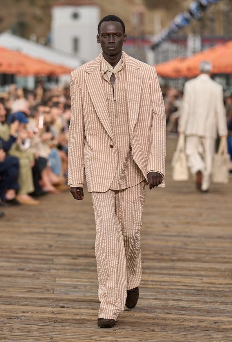Male model wears an oversized tan and cream seersucker suit with peaked lapels over a cream zip-front polo with a brown tie