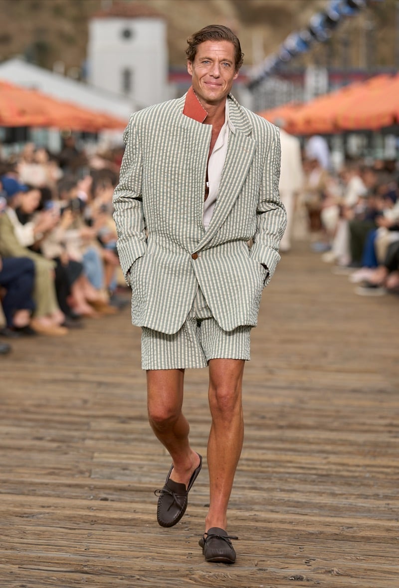 Male model wears a sage green and ivory striped seersucker suit with matching tailored shorts, an open cream shirt, and brown leather moccasin loafers
