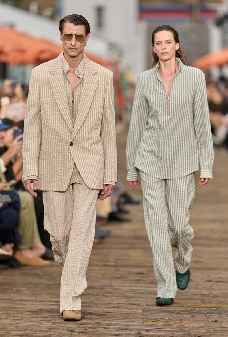 Two male models walk side by side in seersucker suits, one in beige with a half-zip polo, the other in sage and ivory with matching wide-leg trousers