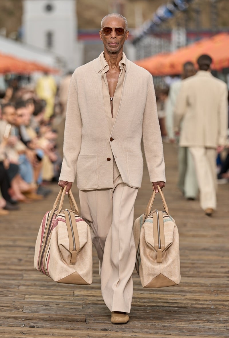 Male model wears a cream boucle knit jacket over a matching half-zip polo and wide-leg ivory trousers, holding two oversized striped canvas duffels