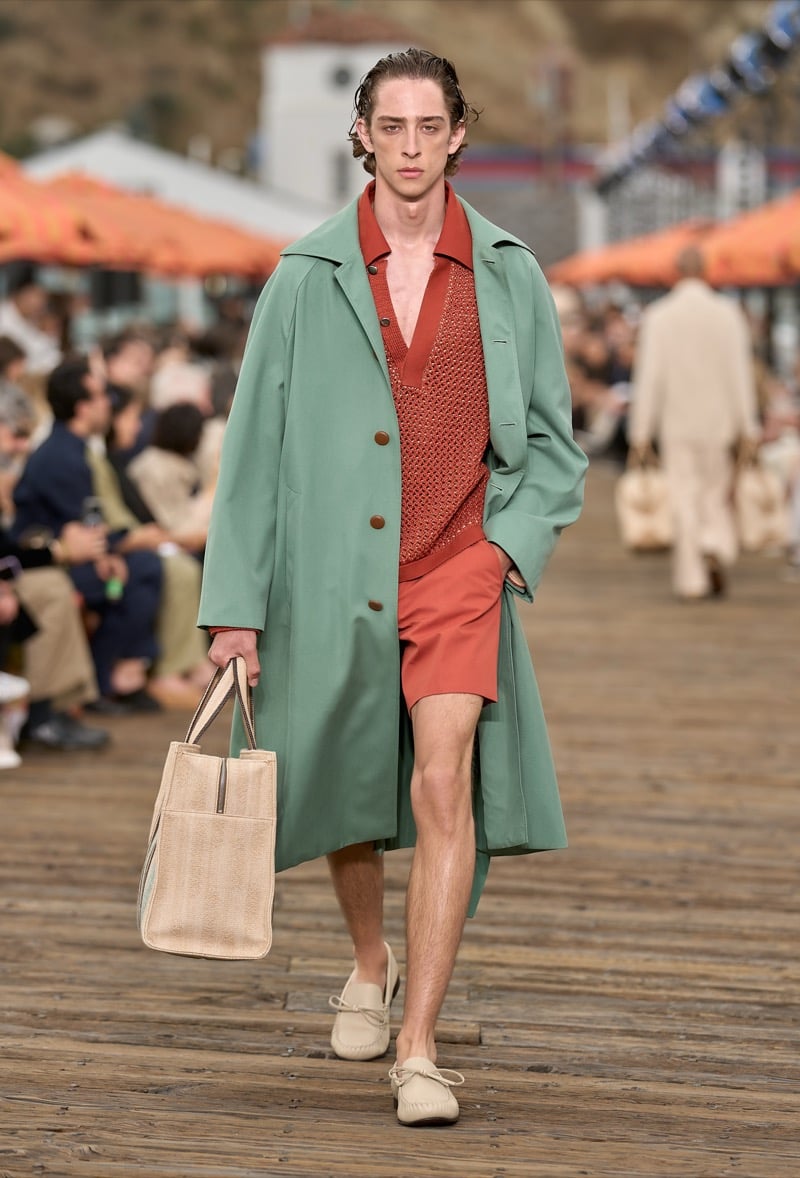 Male model wears a sage green oversized trench coat over a rust knit polo and rust shorts with cream suede driving moccasins, holding a woven straw tote