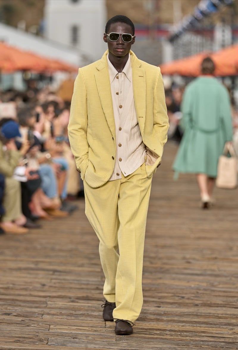 Male model wears a pale yellow textured suit over a cream waistcoat and cream shirt with brown suede shoes and white sunglasses