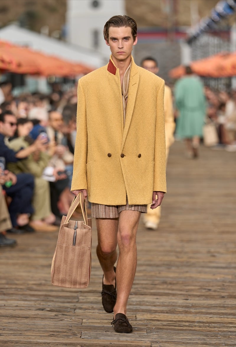Male model wears an oversized butter yellow double-breasted blazer with a brick red collar over brown striped shorts, holding a tan suede tote