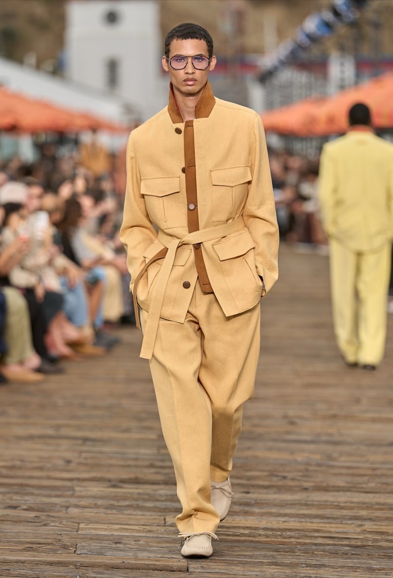 Male model wears a pale yellow belted safari jacket with brown placket detail over matching wide-leg trousers and cream suede moccasins