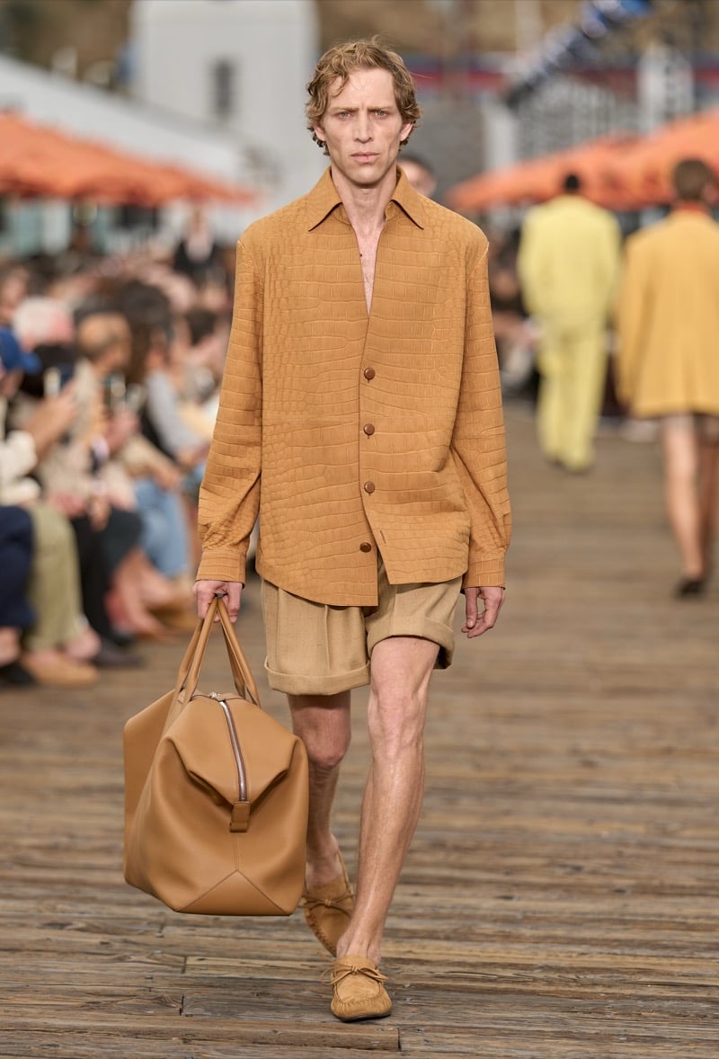 Male model wears a tobacco crocodile-textured leather shirt jacket over tan pleated shorts with tan suede moccasin loafers, carrying a large tan leather weekender