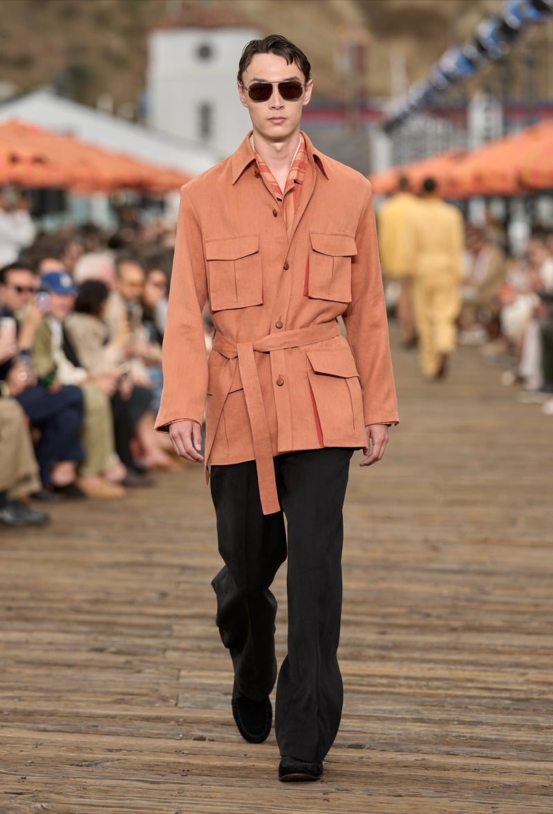 Male model wears a terracotta belted safari overshirt over black wide-leg trousers and black suede moccasins with tortoiseshell sunglasses
