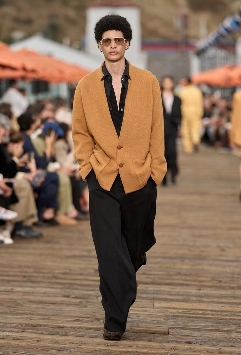 Male model wears a burnt orange ribbed knit cardigan over a black open-collar shirt with black wide-leg trousers and aviator sunglasses