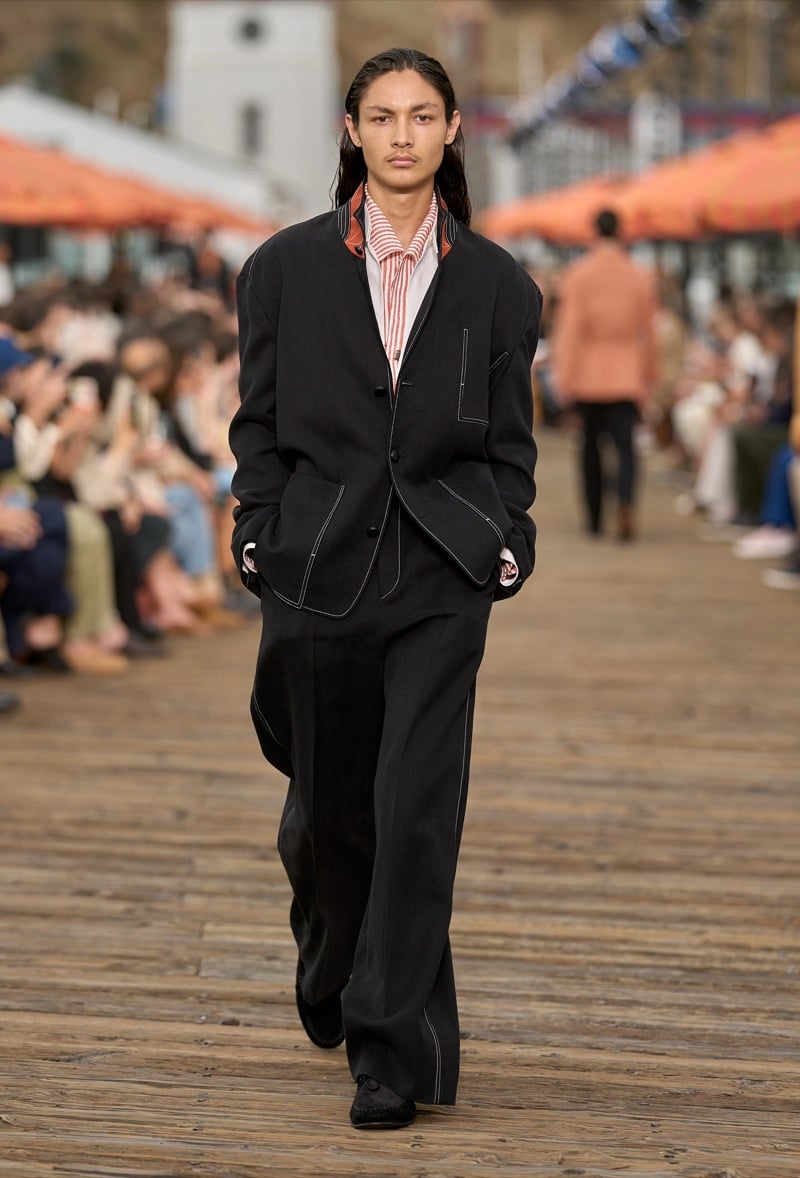 Male model wears an oversized black overshirt with white contrast stitching over a red and white striped shirt and matching wide-leg trousers