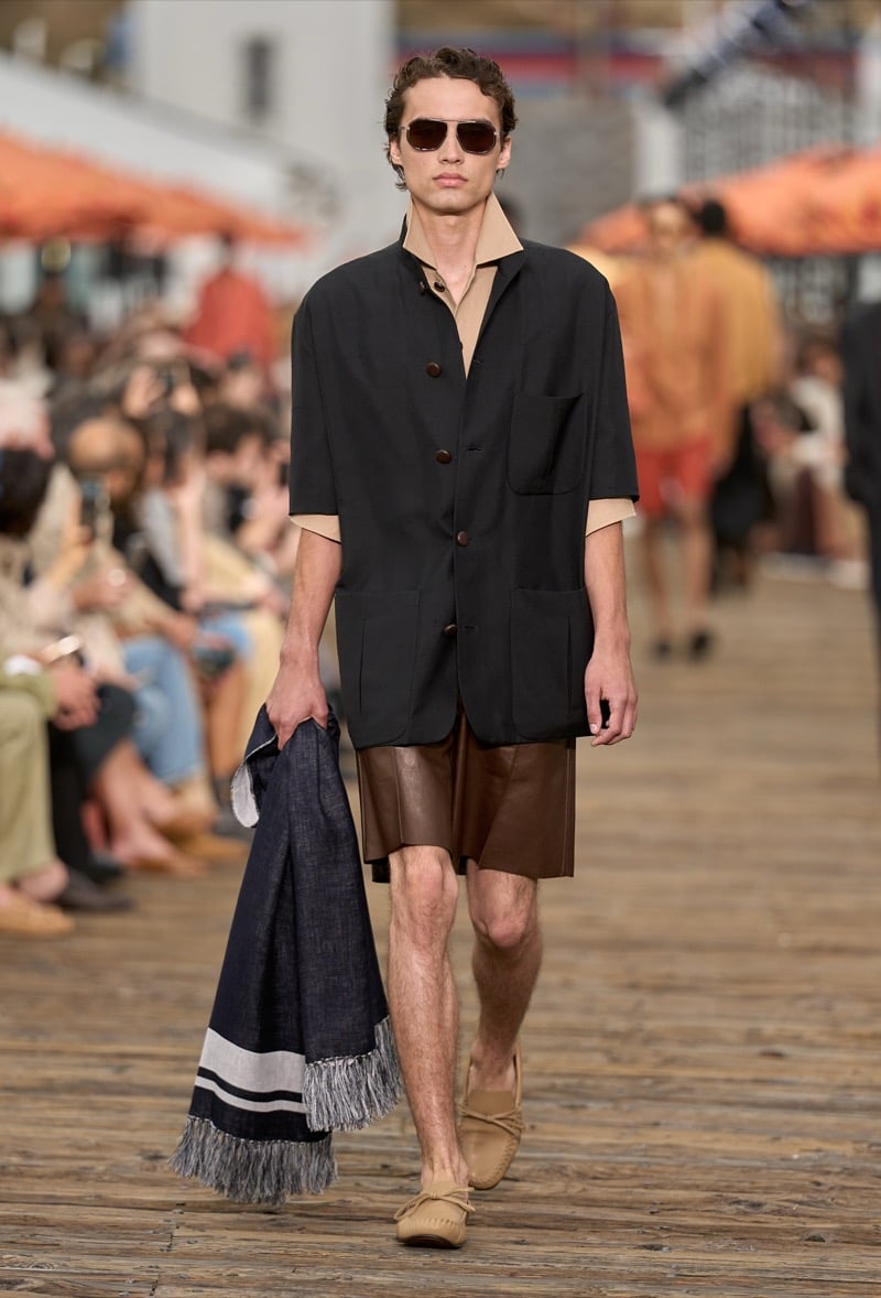 Male model wears a black short-sleeve overshirt over a tan polo with brown leather shorts and tan moccasins, carrying a navy fringed beach towel