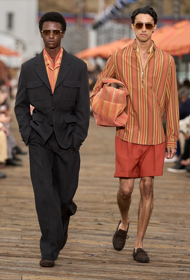 Two male models walk together, one in a charcoal striped suit with an orange striped shirt, the other in a matching orange and rust striped half-zip shirt with rust shorts