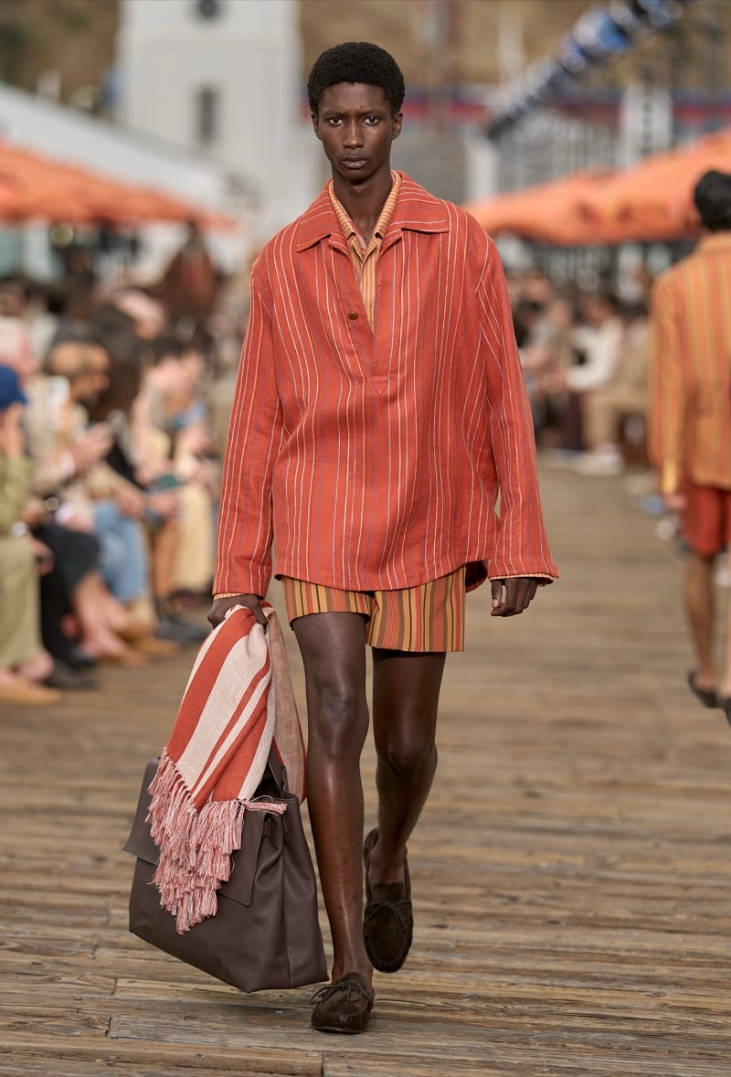 Male model wears a rust and cream pinstripe overshirt over matching striped shorts with brown moccasins, carrying a brown leather tote with a rust striped throw