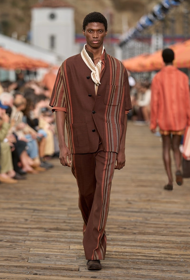 Male model wears a brown and rust irregular striped short-sleeve suit jacket over matching wide-leg trousers with a cream and brown striped silk scarf at the neck