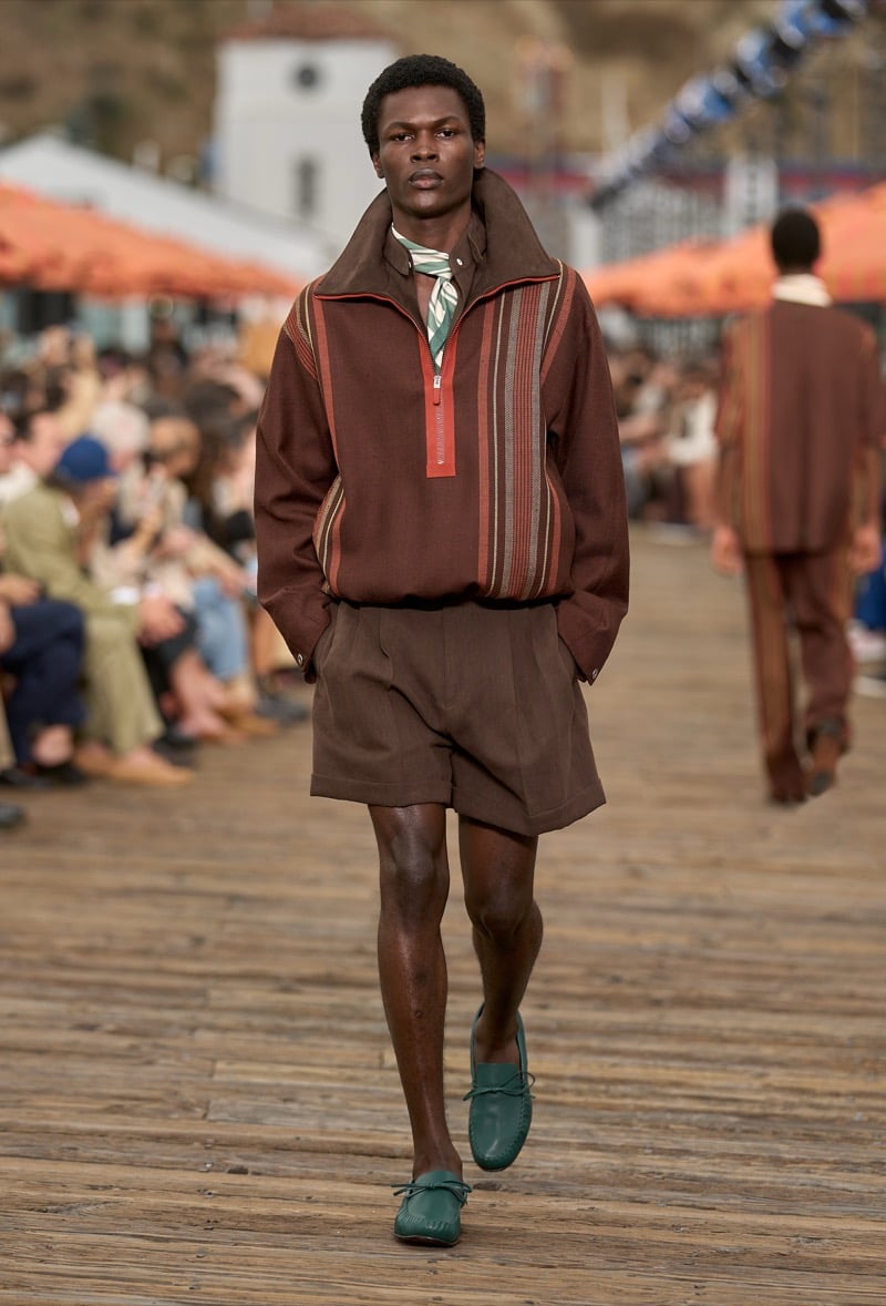 Male model wears a brown striped knitted half-zip pullover with a tall collar over matching brown pleated shorts with green suede moccasin loafers