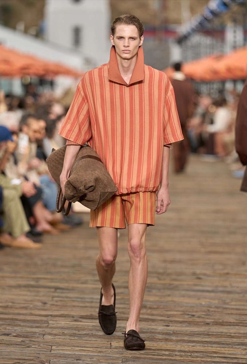 Male model wears an oversized rust and orange striped knit polo with a tall collar over matching striped shorts, holding a brown croc-textured tote