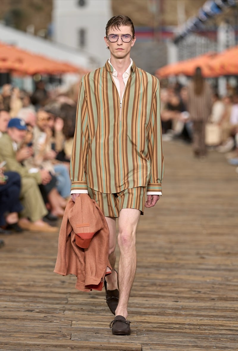Male model wears a green and rust striped silk pajama set with matching shorts, carrying a folded rust knit blazer