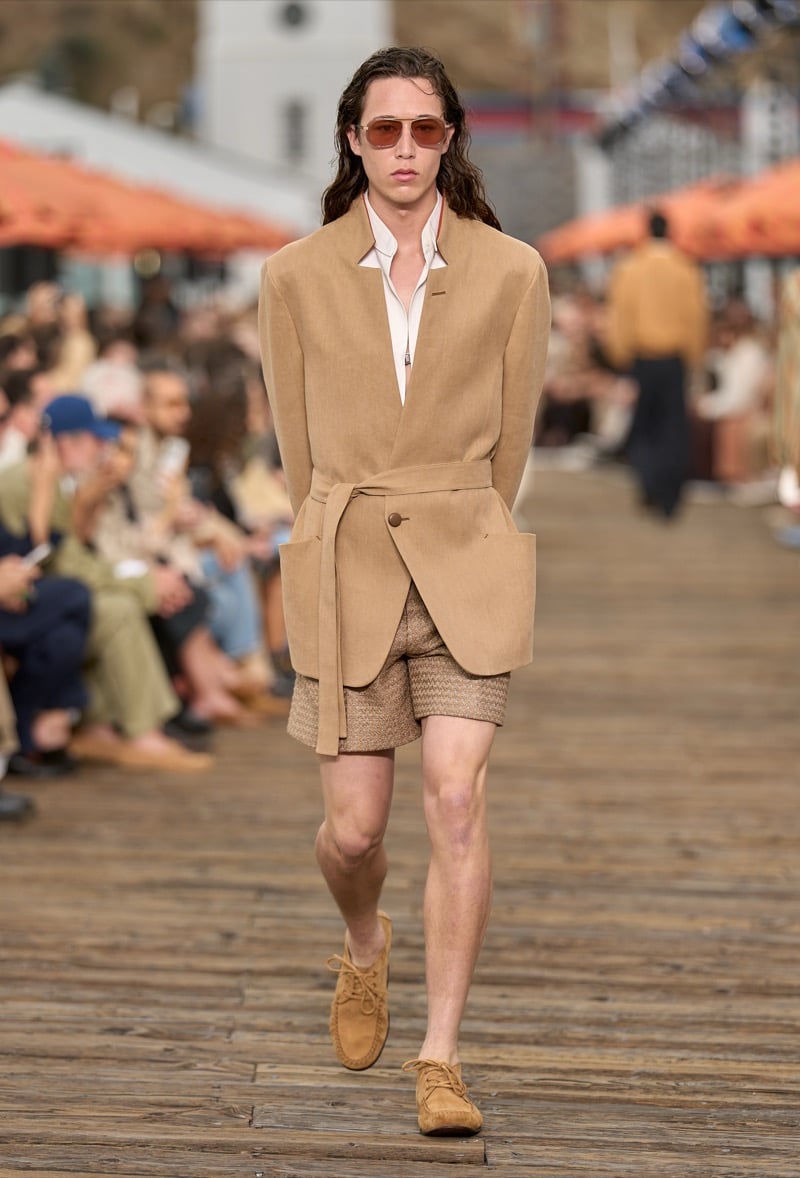 Male model wears a camel linen belted wrap jacket with stand collar over woven tan shorts and tan suede lace-up moccasins