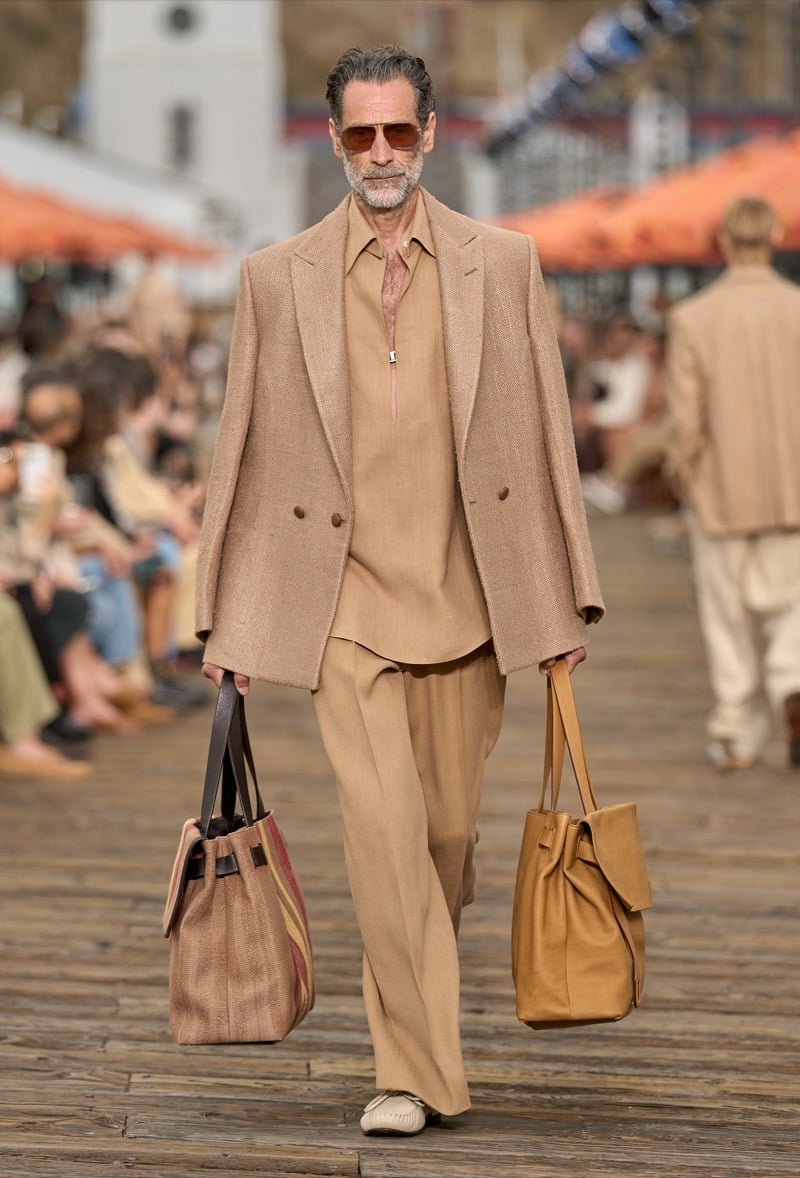 Male model wears a camel textured double-breasted blazer over a tan half-zip silk shirt and matching wide-leg trousers, carrying two oversized leather totes