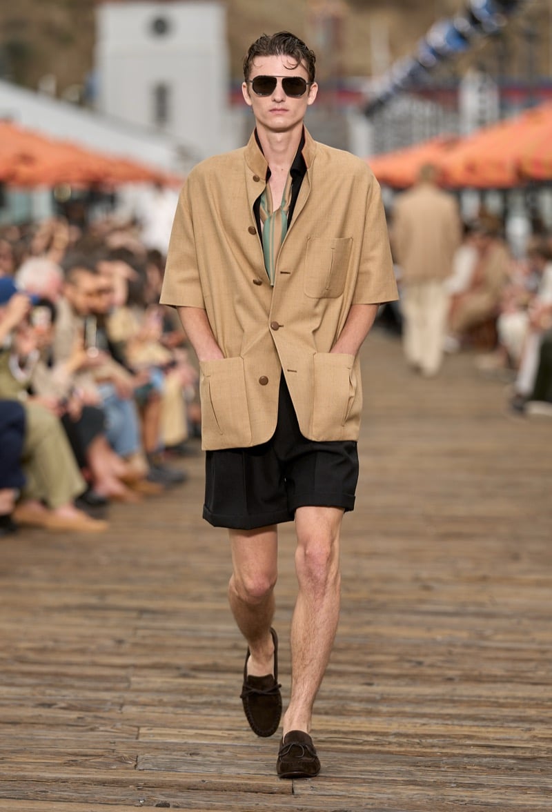 Male model wears a camel short-sleeve safari jacket over a sage and green striped shirt with black tailored shorts and brown suede moccasins