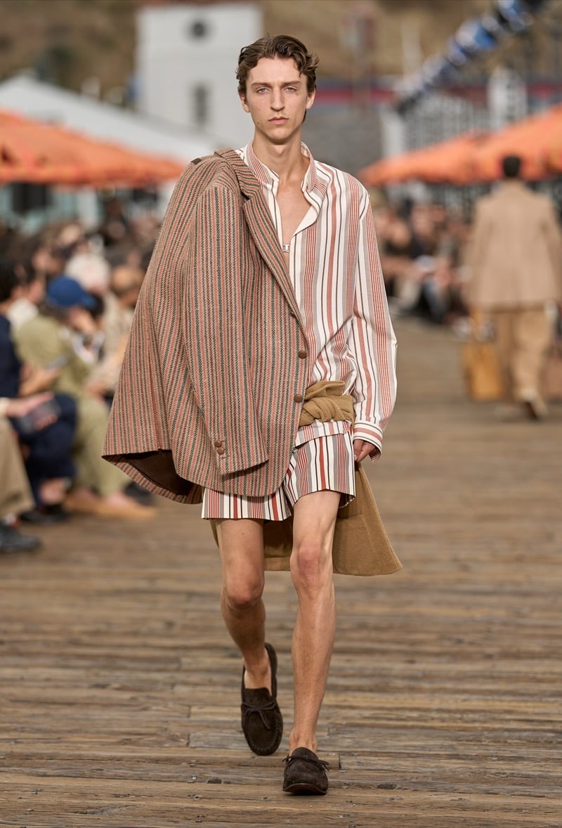 Male model wears a brown ribbed knit jacket draped over the shoulders with a cream and rust striped pajama shirt and matching shorts