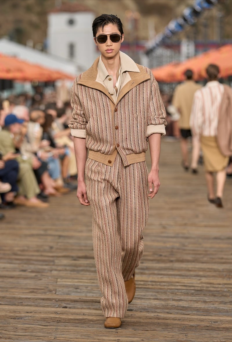 Male model wears a tan striped knit two-piece set with a high-collar zip-front pullover and matching pleated wide-leg trousers with tan suede shoes