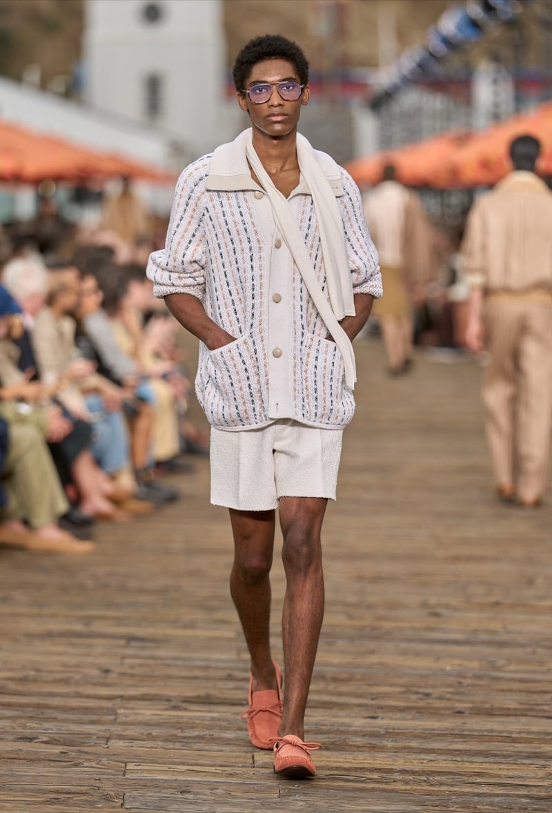 Male model wears a cream textured woven cardigan with multicolor flecks over matching cream toweling shorts with rust suede moccasins, a cream knit draped at the shoulders
