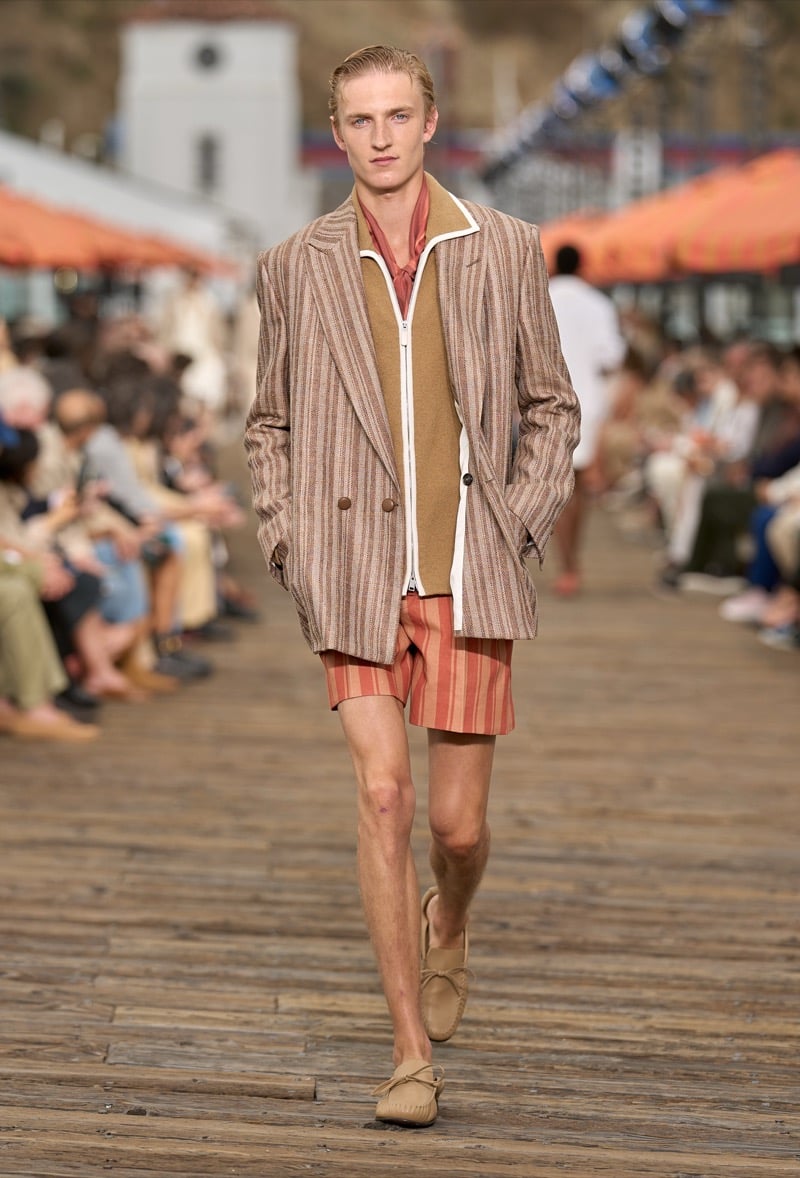 Male model wears a brown and rust pinstripe double-breasted blazer over a tan knit zip-front polo with rust and cream striped shorts and tan moccasins