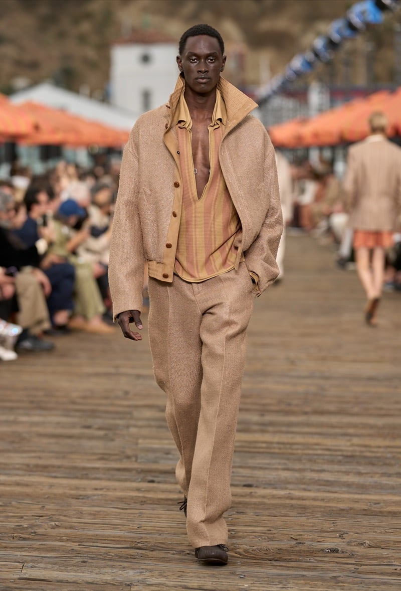 Male model wears a tan textured bomber jacket over a yellow and rust striped silk shirt with matching tan textured wide-leg trousers