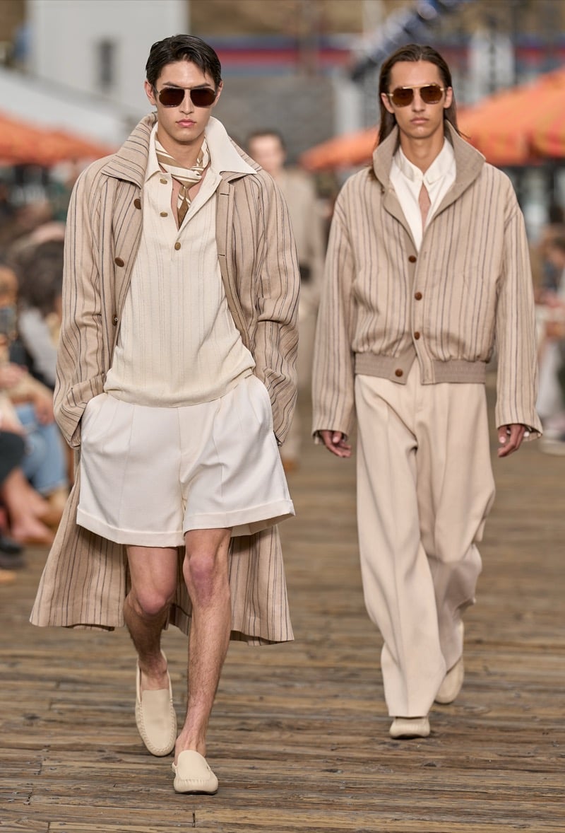 Two male models walk together, one in a tan pinstripe trench over a cream knit polo and white pleated shorts, the other in a tan striped knit cardigan with cream wide-leg trousers