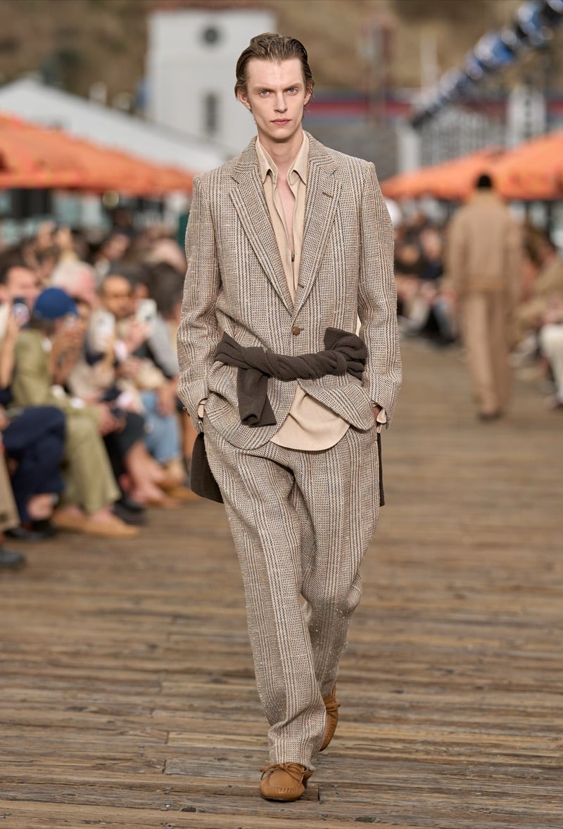 Male model wears a beige herringbone striped single-breasted suit over a pink half-zip shirt with a brown knit tied at the waist and tan suede moccasins