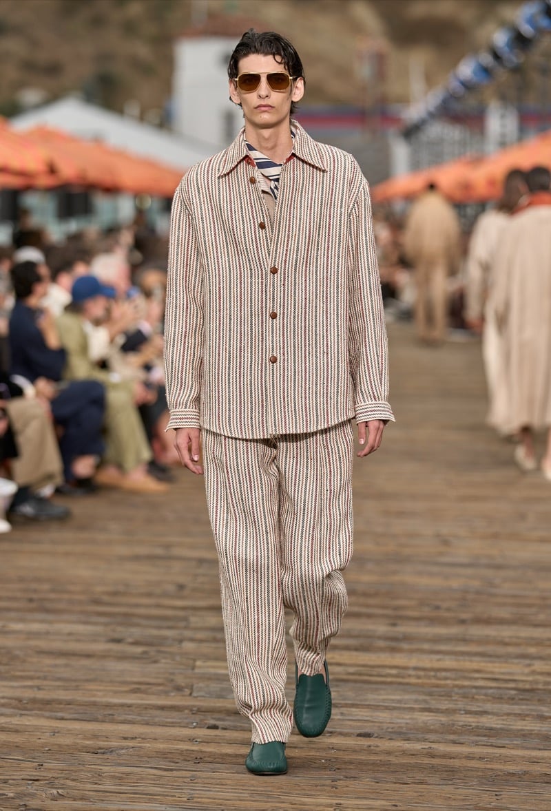 Male model wears a cream and rust pinstripe overshirt over matching wide-leg trousers with a striped collar layer underneath and green suede moccasins