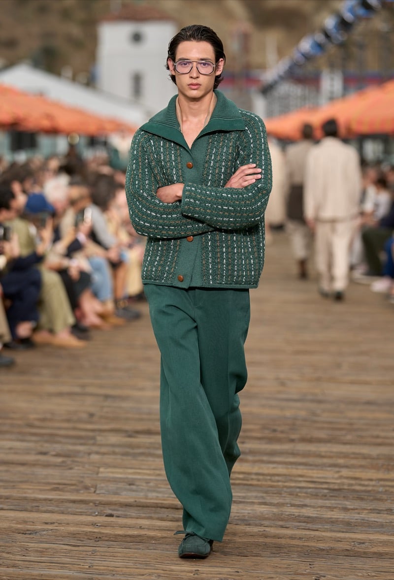 Male model wears a forest green and white striped knit cardigan over matching forest green wide-leg trousers with green suede shoes and clear blue glasses