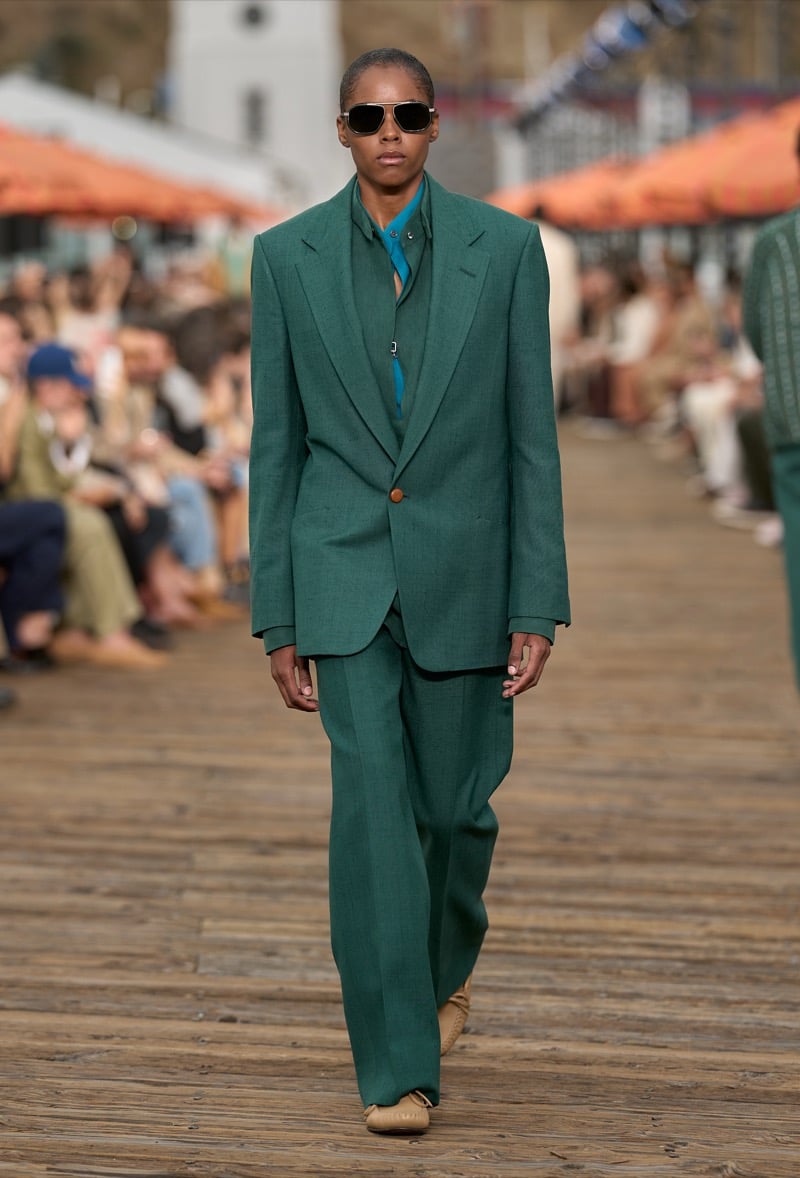 Male model wears an oversized emerald green peak lapel single-breasted suit over a teal half-zip shirt with tan suede moccasins and aviator sunglasses