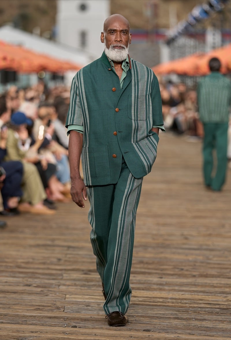 Male model wears a forest green and pale green vertical stripe short-sleeve suit over matching wide-leg trousers with brown suede moccasins