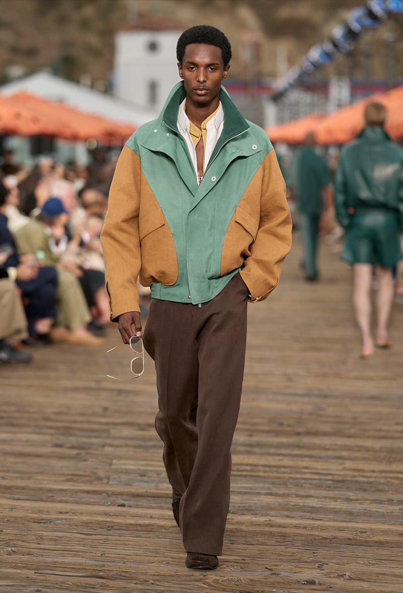 Male model wears a sage green and tobacco color-blocked nappa leather bomber jacket over a cream shirt with brown wide-leg trousers, holding white sunglasses