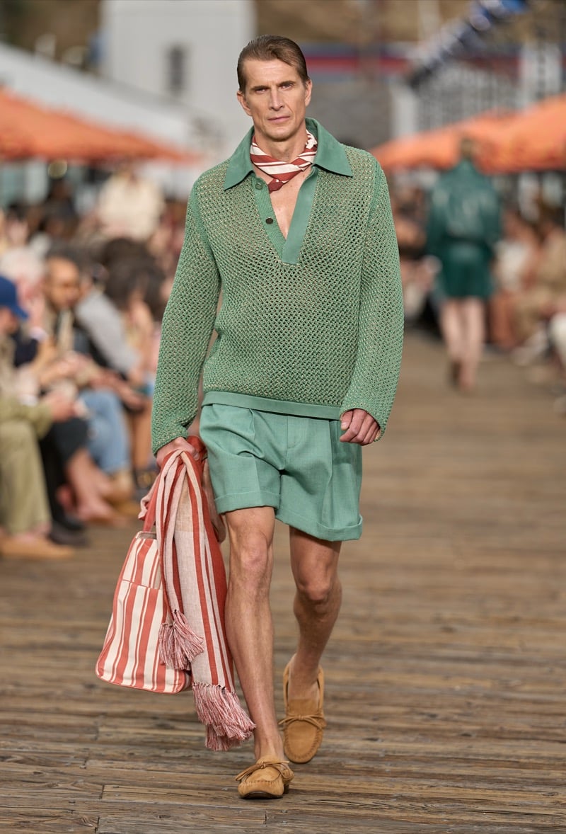 Male model wears a sage green open knit mesh polo over matching pleated shorts with tan suede moccasins, carrying a rust and white striped beach towel and tote