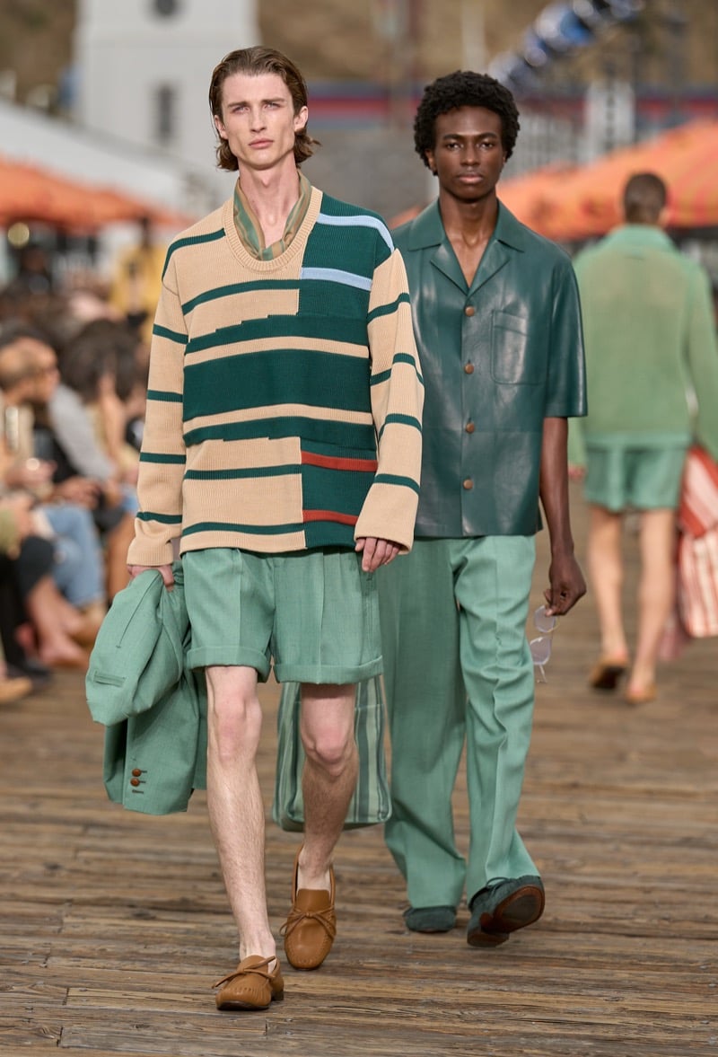 Two male models walk together, one in a cream and green striped patchwork knit with sage shorts, the other in a teal nappa leather camp shirt with sage wide-leg trousers