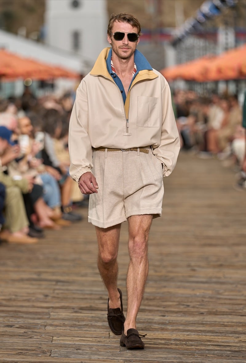 Male model wears a cream half-zip windbreaker with yellow and blue collar accents over linen pleated shorts with a woven belt and dark brown suede moccasins