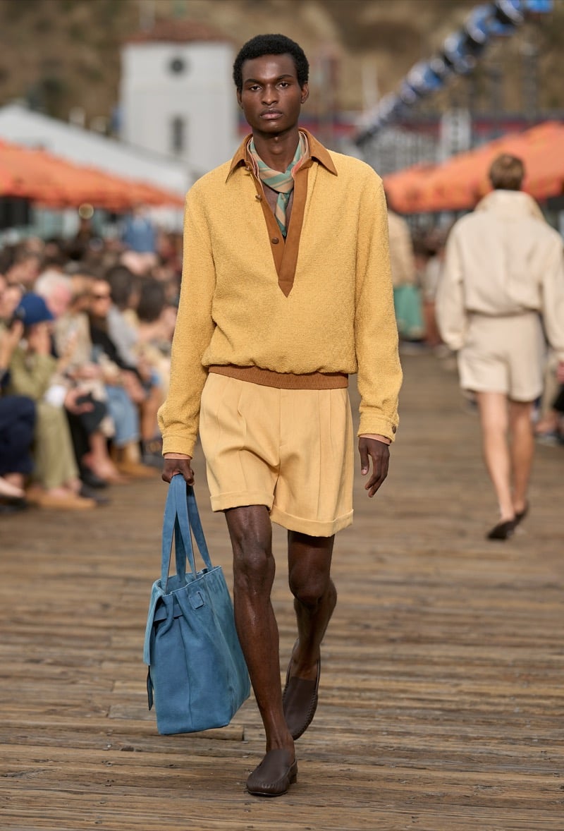 Male model wears a butter yellow textured half-zip pullover over tobacco-collared layer and matching yellow pleated shorts with brown moccasins, carrying a blue suede tote